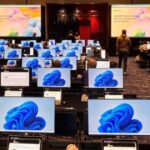 Ein Konferenzraum mit sehr vielen Arbeitsplätzen; einige Menschen sitzen an den Arbeitsplätzen und schauen auf die Monitore